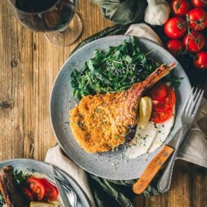 A wooden table with two plates of golden brown veal milanese, greens, and sliced tomatoes.