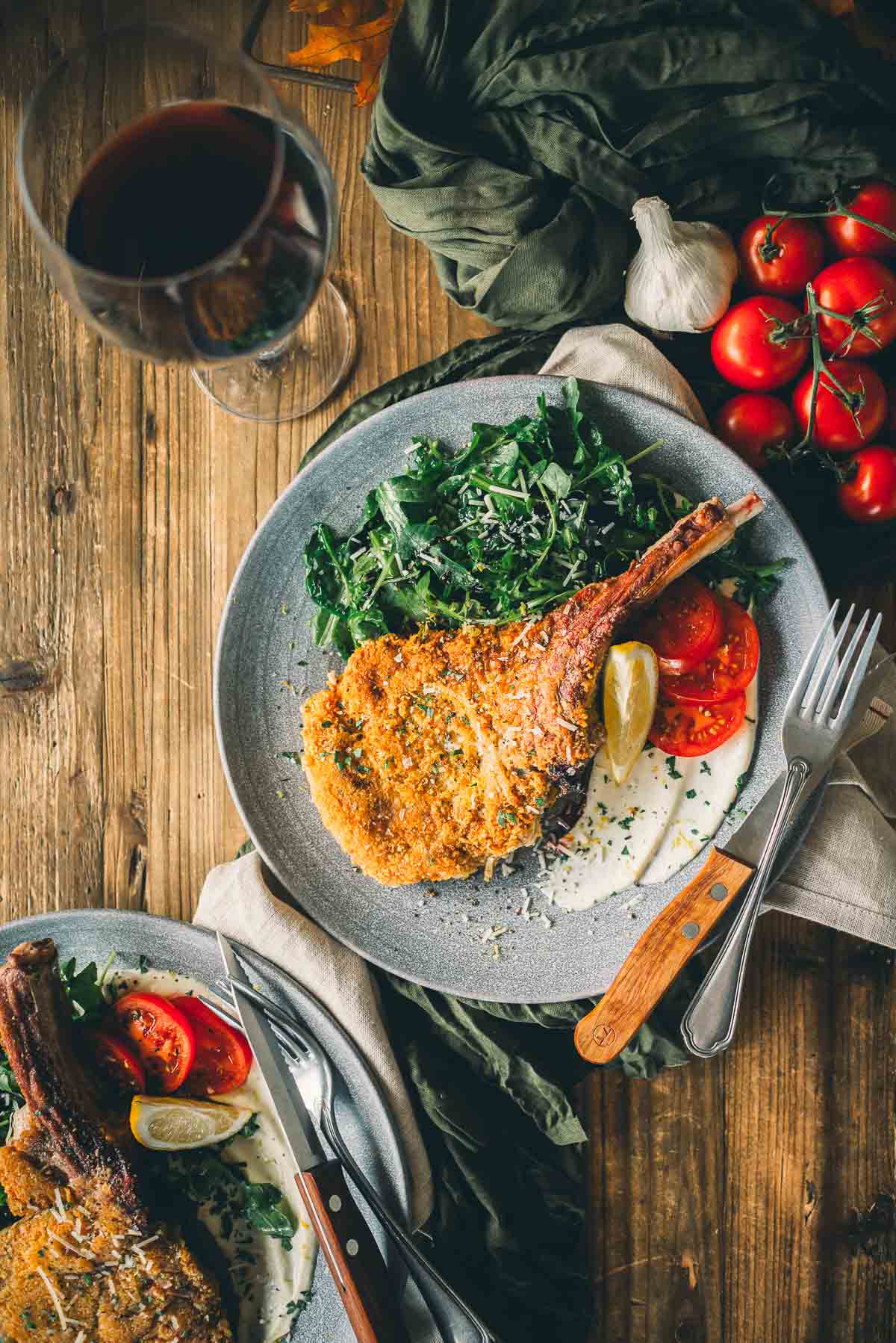 A wooden table with two plates of golden brown veal milanese, greens, and sliced tomatoes. 