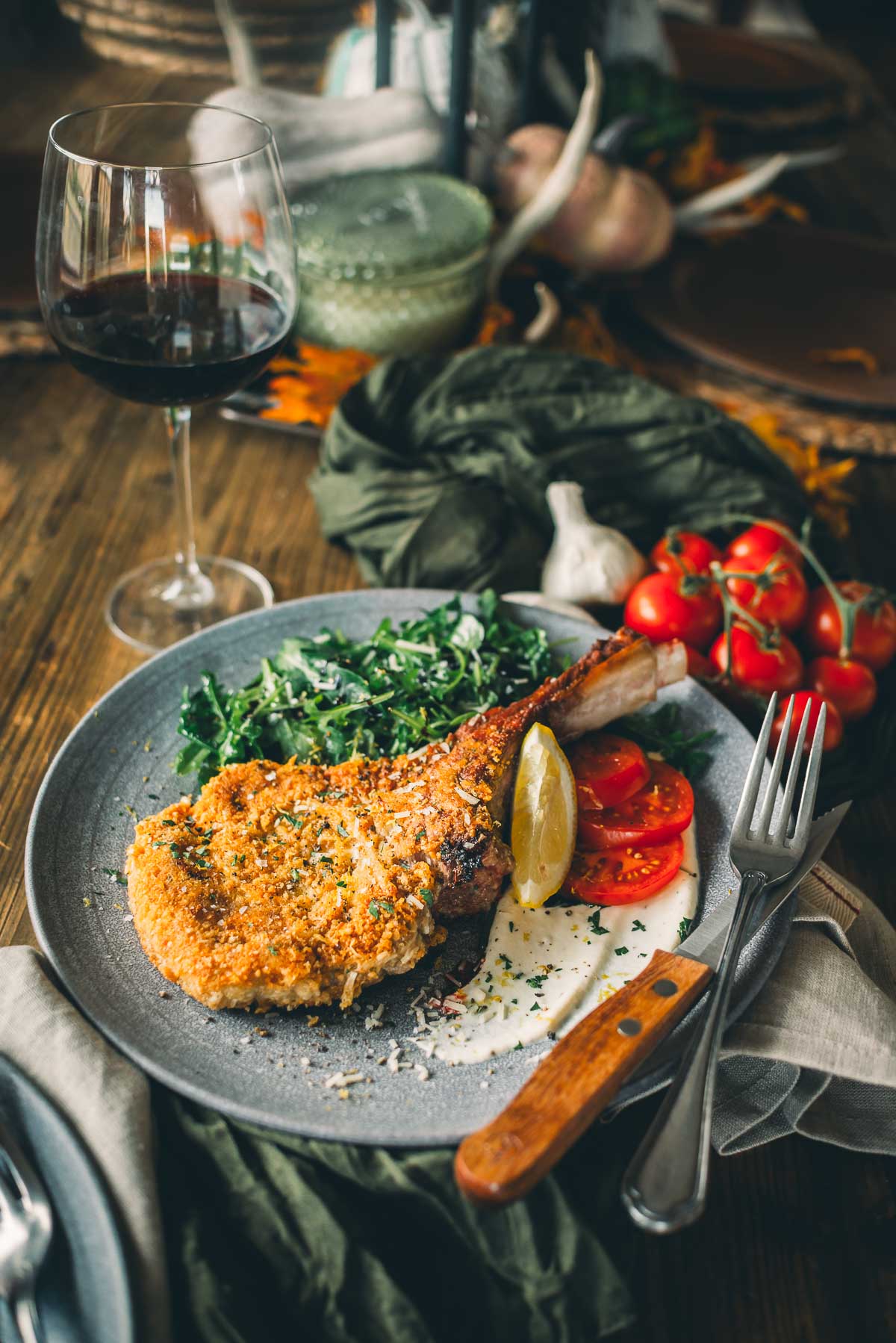 A plated meal featuring a breaded veal milanese with a lemon wedge, sliced tomatoes, leafy greens, and a glass of red wine on a wooden table.