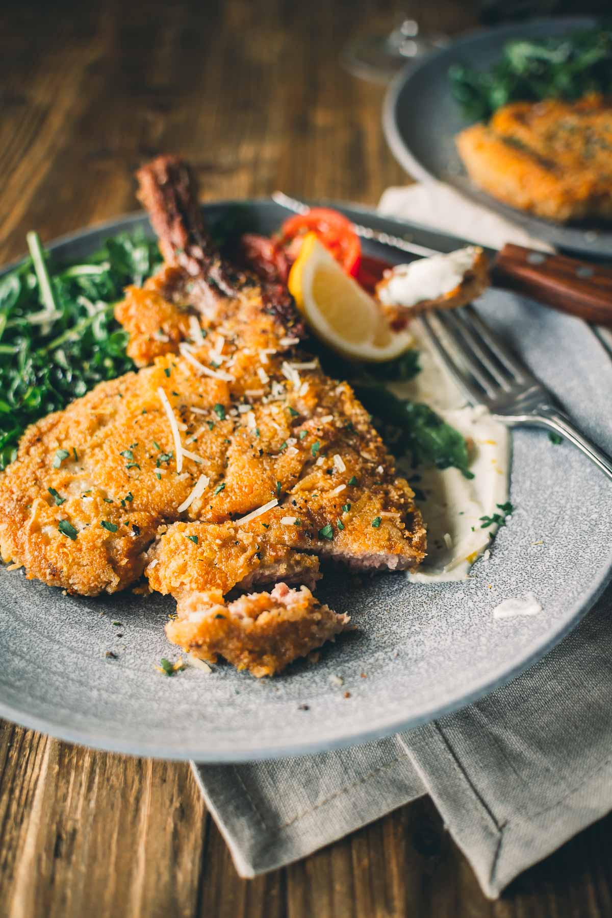 A plate of crispy breaded veal chop served with greens, cherry tomatoes, lemon wedge, and garnished with grated cheese on a wooden table. 