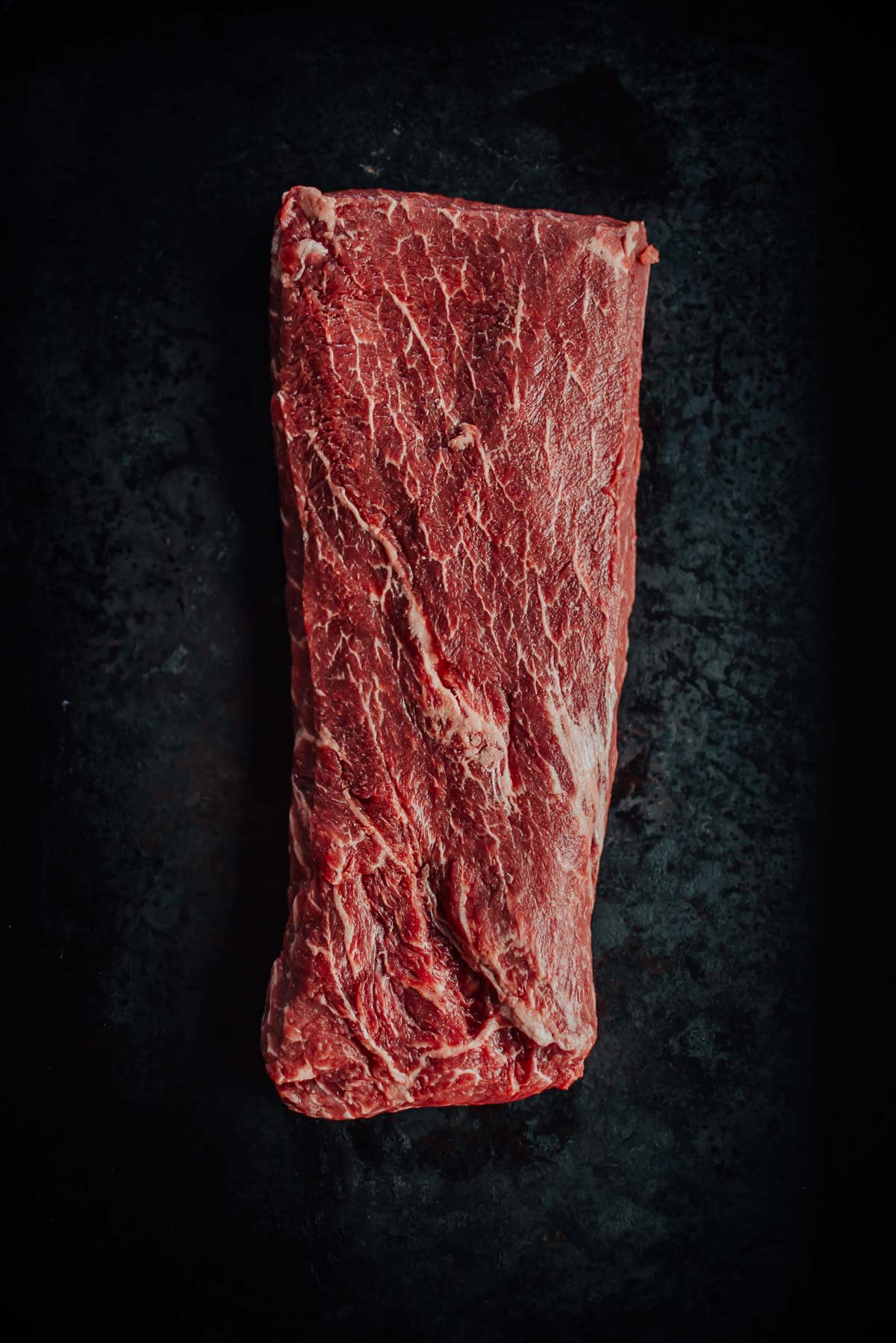 A raw, flat iron steaks sits against a dark background.