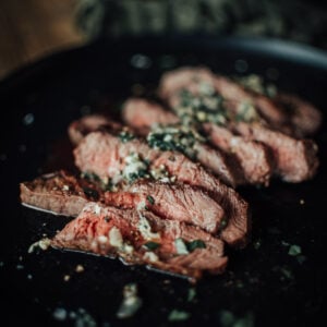 Sliced medium-rare steak with herb butter on a black plate.