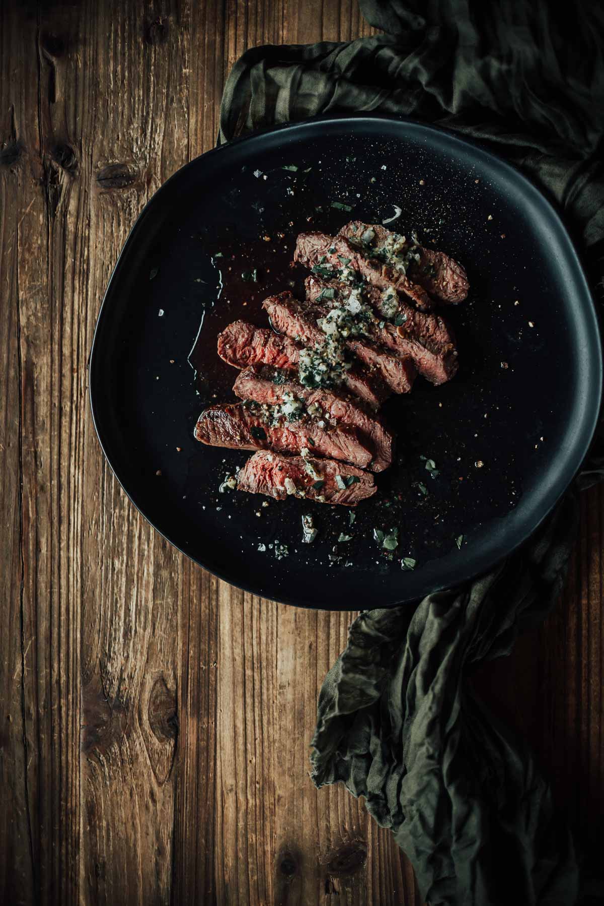 A black plate on a wooden table holds sliced, medium-rare flat iron steak garnished with herbs and seasoning. A green fabric is casually draped beside the plate.