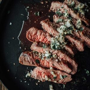 A plate of sliced flat iron steak topped with herbs and seasoning, served on a dark dish.