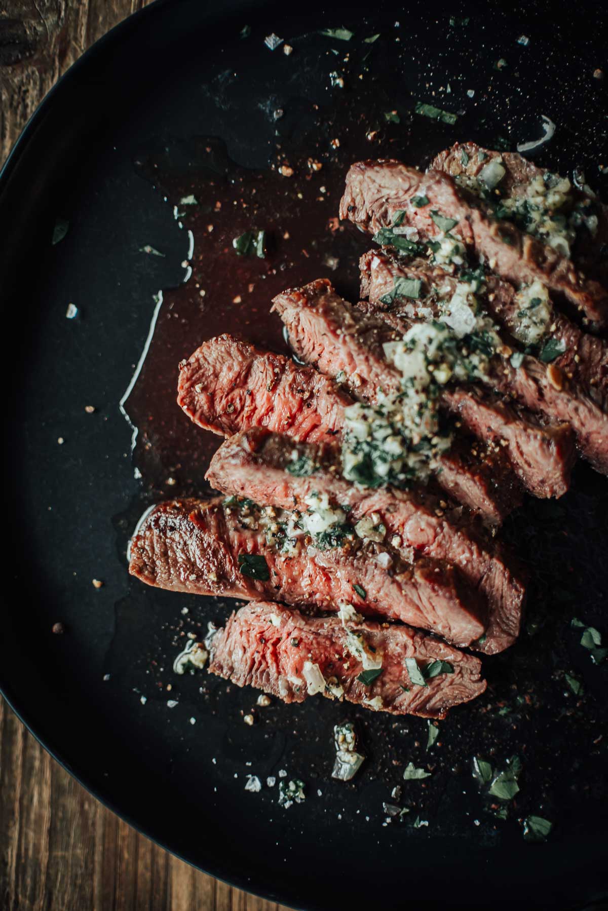 A plate of sliced flat iron steak topped with herbs and seasoning, served on a dark dish.