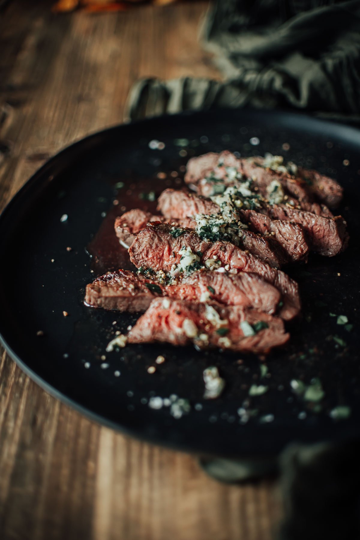 Slices of medium-rare flat iron steak topped with herbs and salt are served on a black plate, placed on a wooden table.