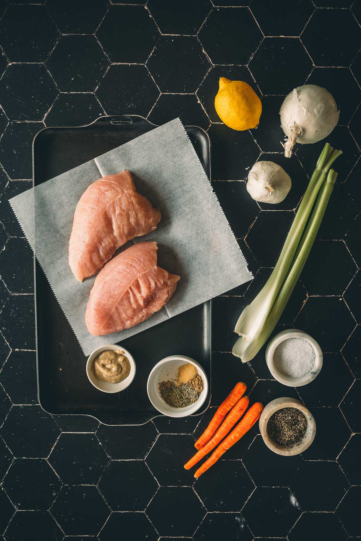 Raw turkey tenderloins on parchment paper in a baking pan surrounded by carrots, celery, onion, a lemon, garlic, and small bowls of mustard, herbs, and spices, all placed on a black tile surface.