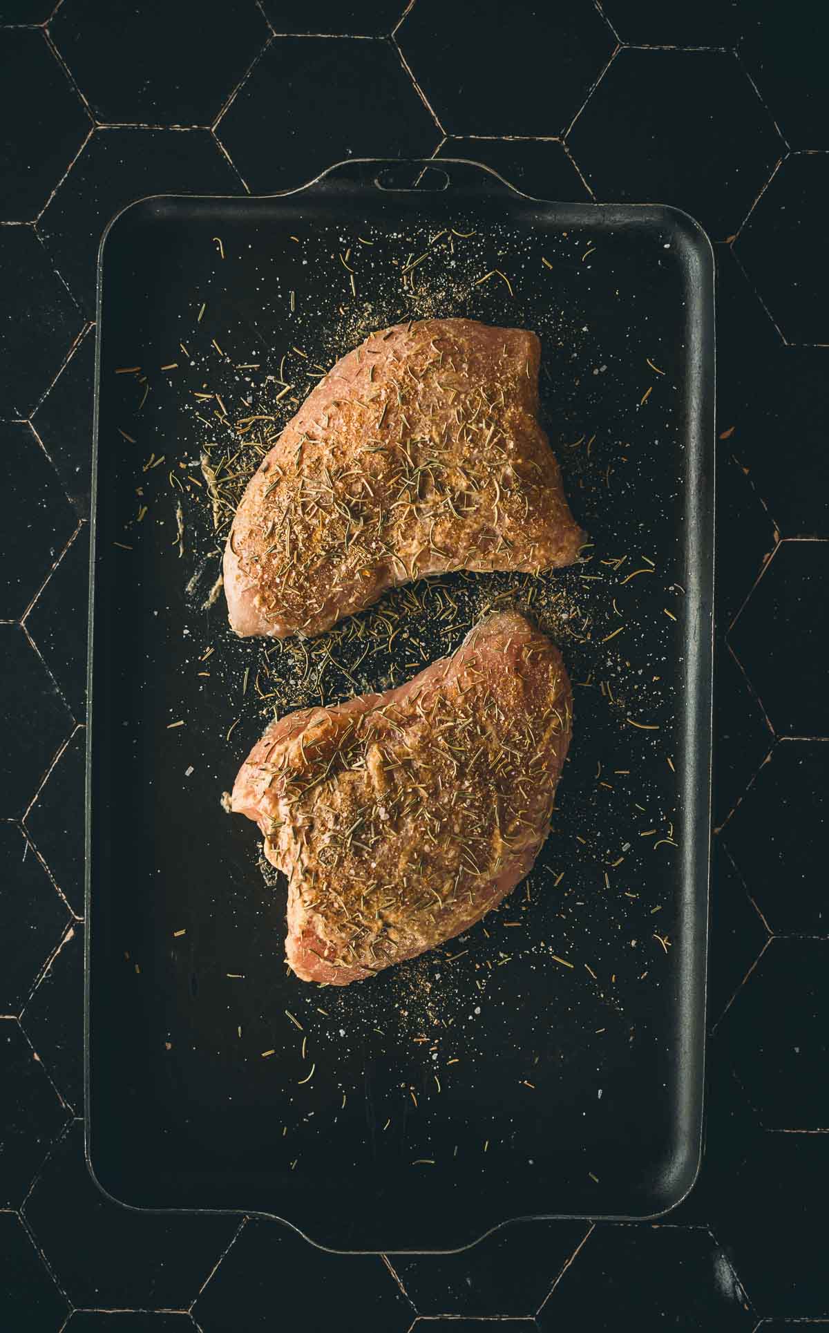 Two seasoned raw turkey tenderloins coated in herbs on a black baking tray, ready for cooking. The tray is placed on a black tile surface.