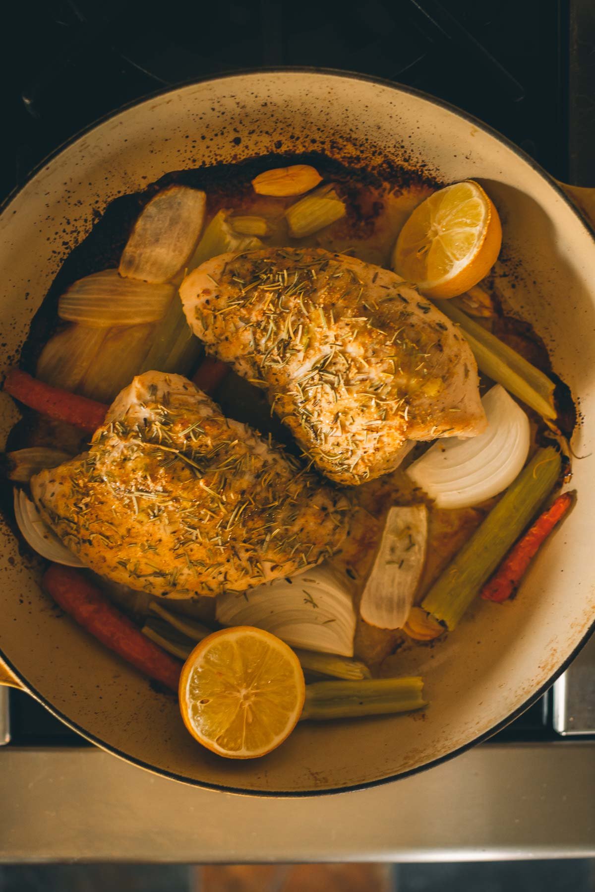 A pan with cooked turkey tenderloins seasoned with herbs, accompanied by onion slices, carrot sticks, celery stalks, garlic cloves, and halved lemon.