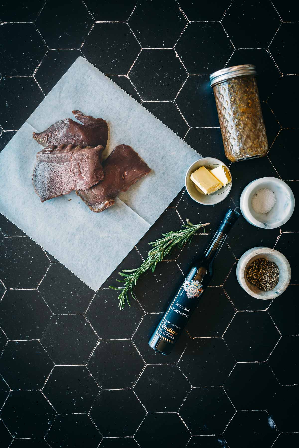 Raw elk steaks on a parchment paper, jar of seasoning, bowl of butter, rosemary, balsamic vinegar bottle, and bowls of salt and pepper placed on a black tiled surface.