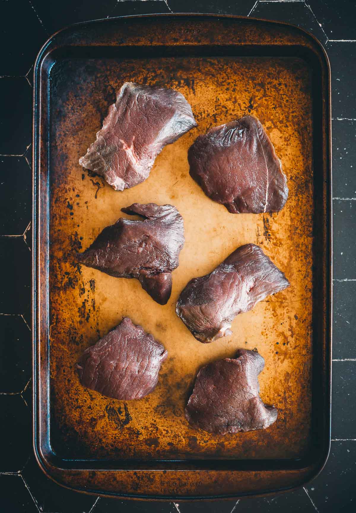 Six raw elk steaks displayed on a well-used, darkened baking sheet.