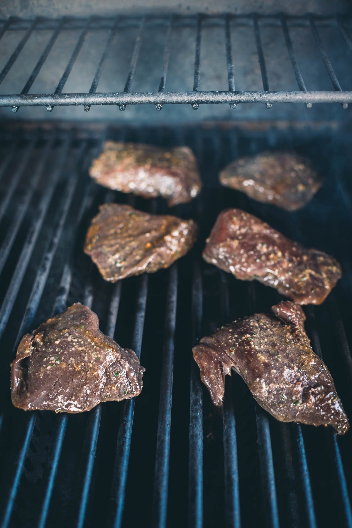 Six pieces of marinated elk steaks being grilled on a barbecue with visible grill marks.