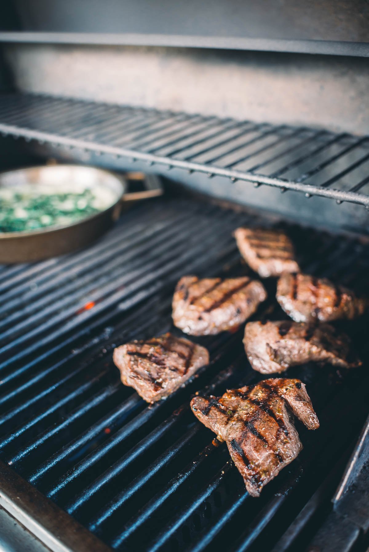 Several elk steaks are grilling on a barbecue. A skillet with vegetables is also visible in the background.