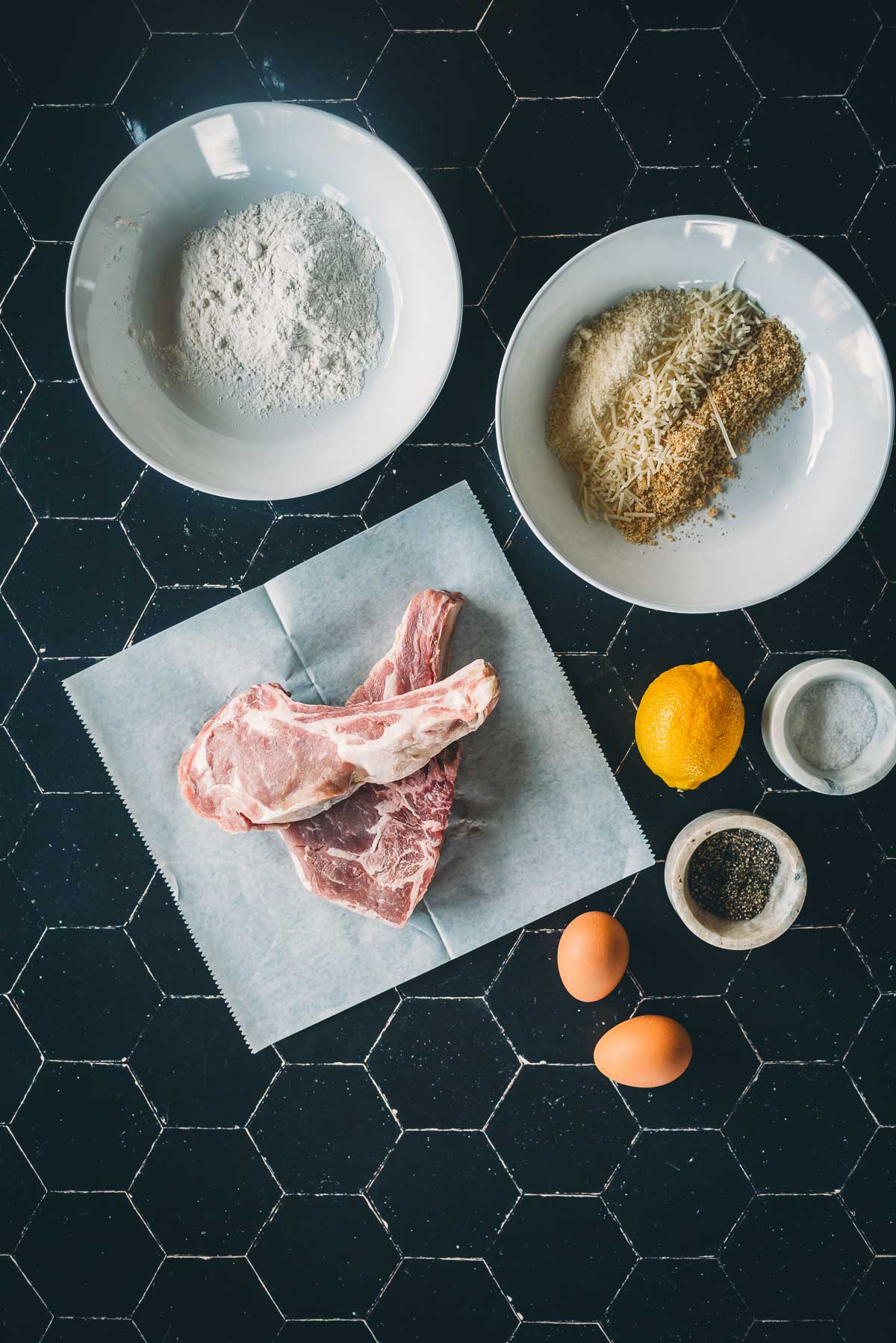 Ingredients for cooking: raw veal chops on paper, a bowl of flour, a bowl with breadcrumbs and grated cheese, lemon, salt, pepper, two eggs on a black tiled surface.