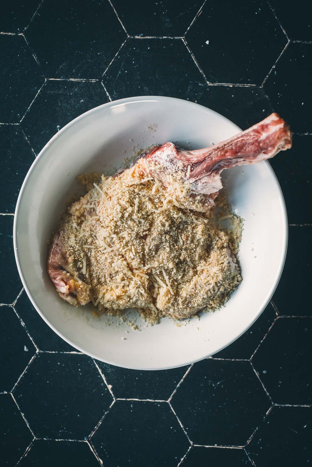 A raw bone-in veal chop in a white bowl to be coated in breading.