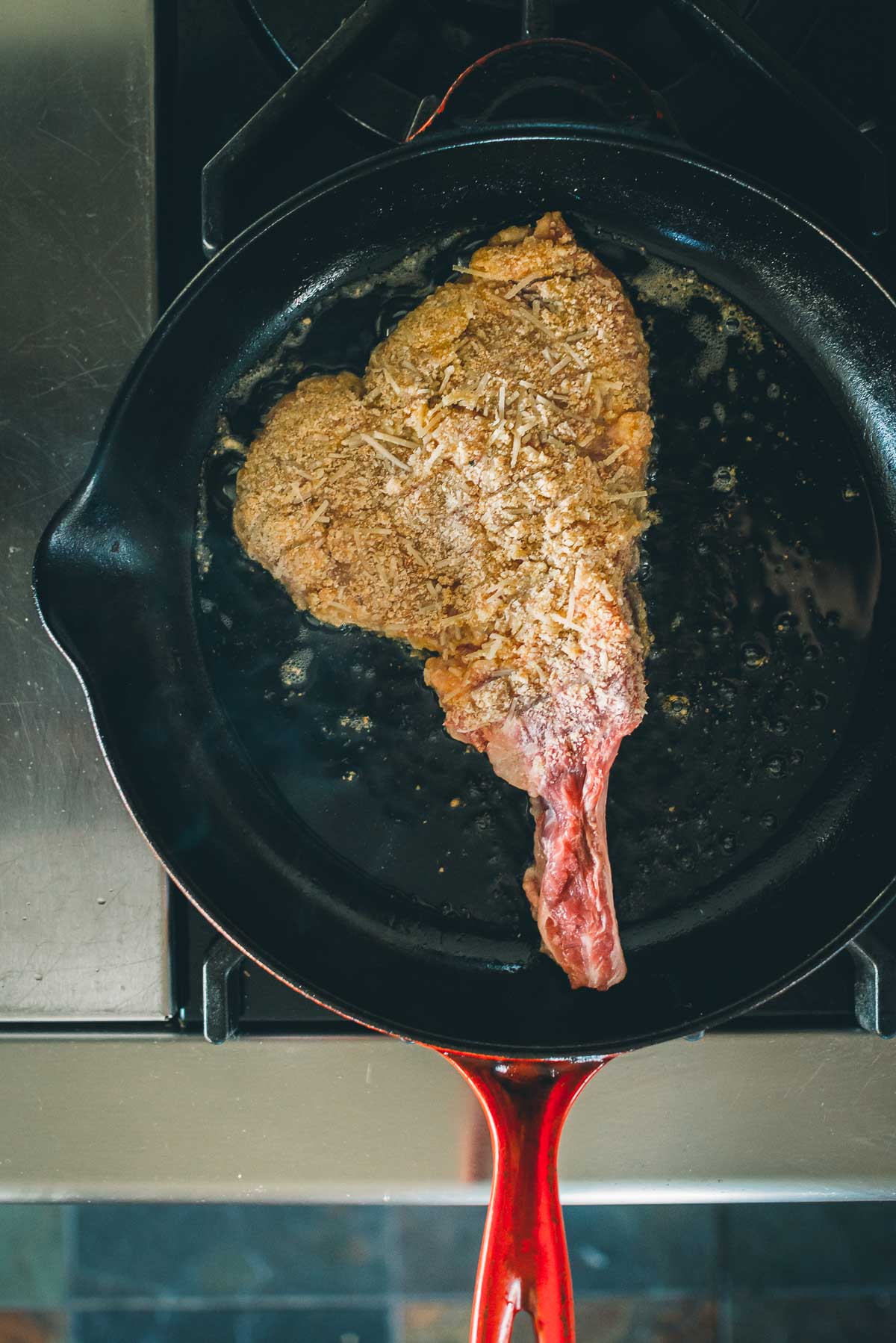 A breaded veal chop is being cooked in a cast iron skillet on a stovetop.