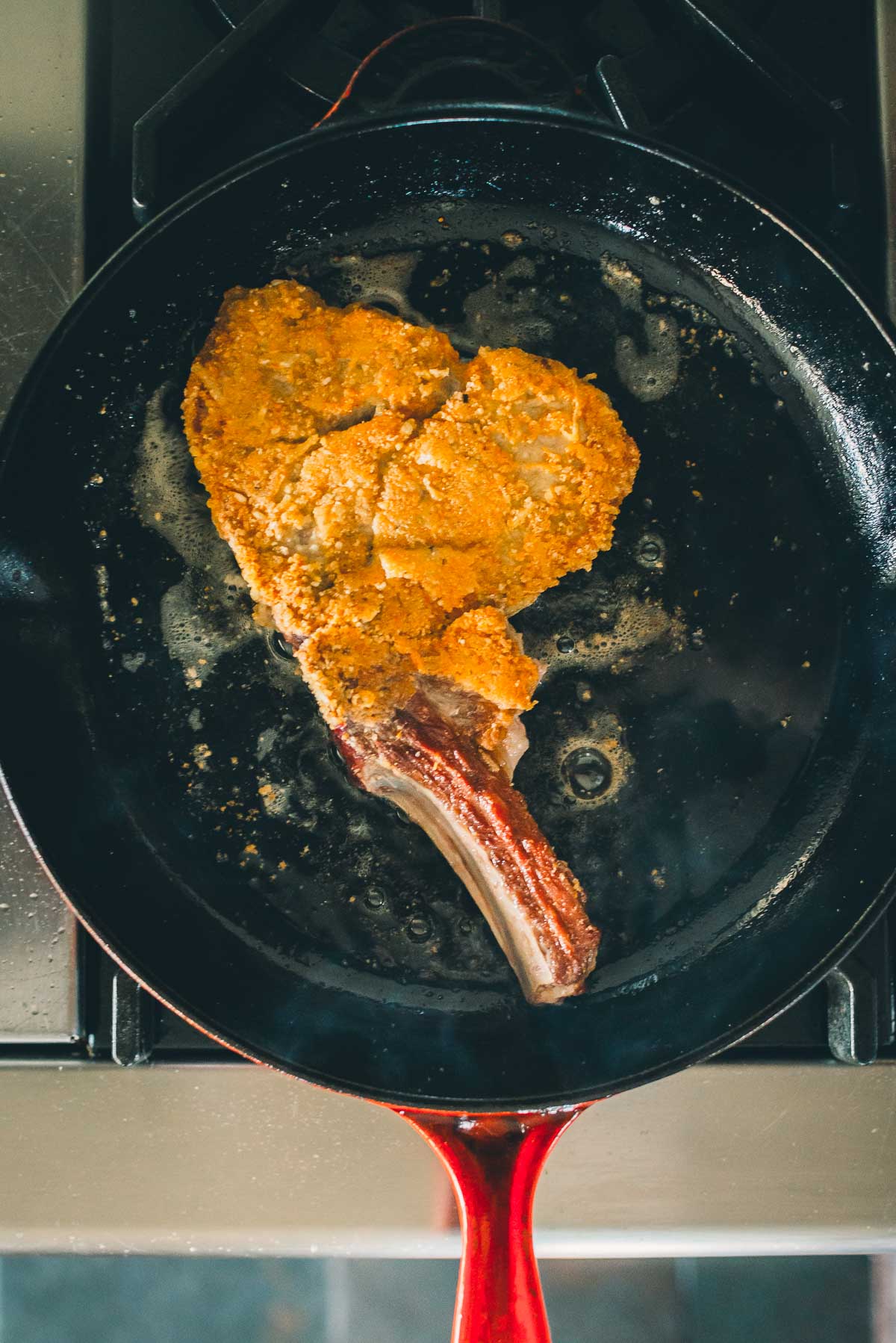 A seasoned, veal chop is being fried in a red-handled black skillet on a stove.