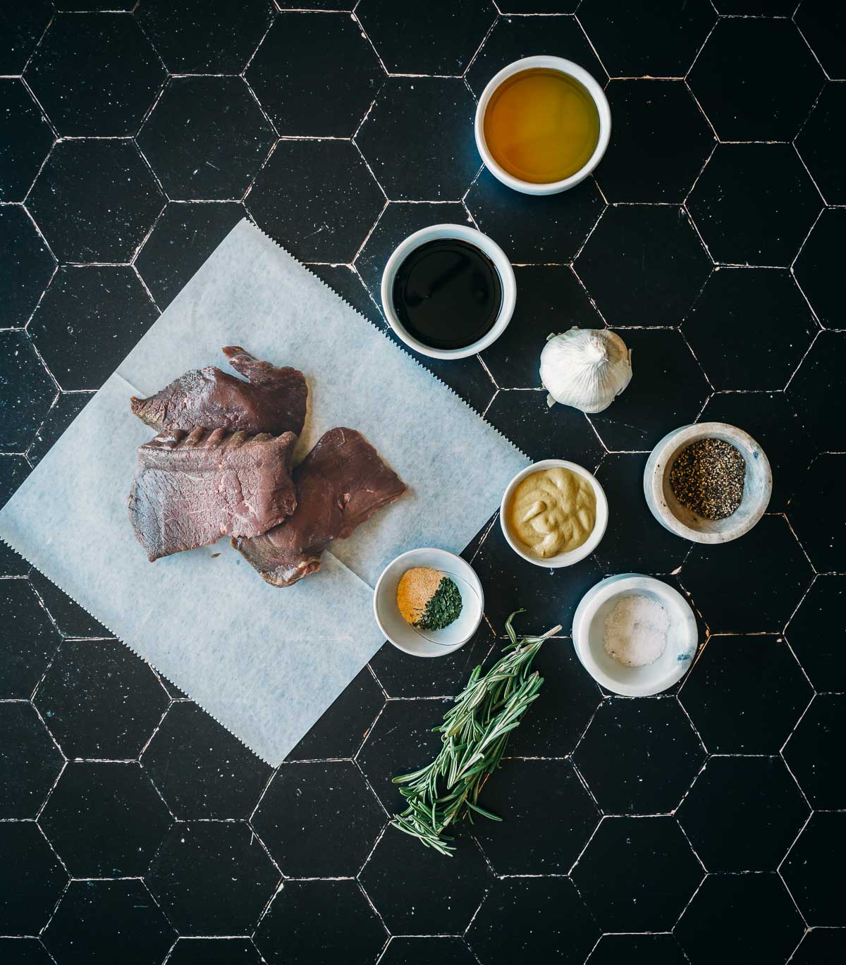 Overhead view of elk steaks on parchment paper surrounded by small bowls containing oil, soy sauce, garlic, mustard, pepper, salt, an herb sprig, and a mix of seasonings, on a black surface.