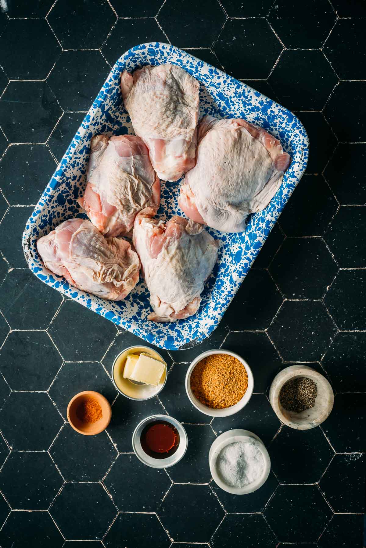 Five raw turkey thighs in a blue speckled tray, surrounded by small bowls containing various seasonings and ingredients on a black tile surface.