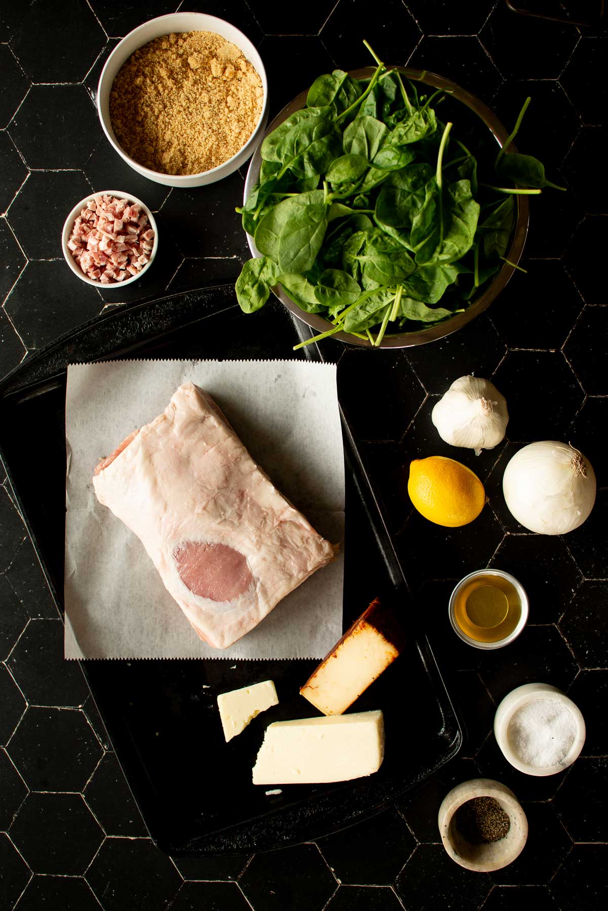 Overhead view of cooking ingredients including spinach, minced panchetta, brown sugar, a piece of raw pork loin on parchment paper, cheese, garlic, lemon, onion, and small bowls of oil, salt, and pepper.