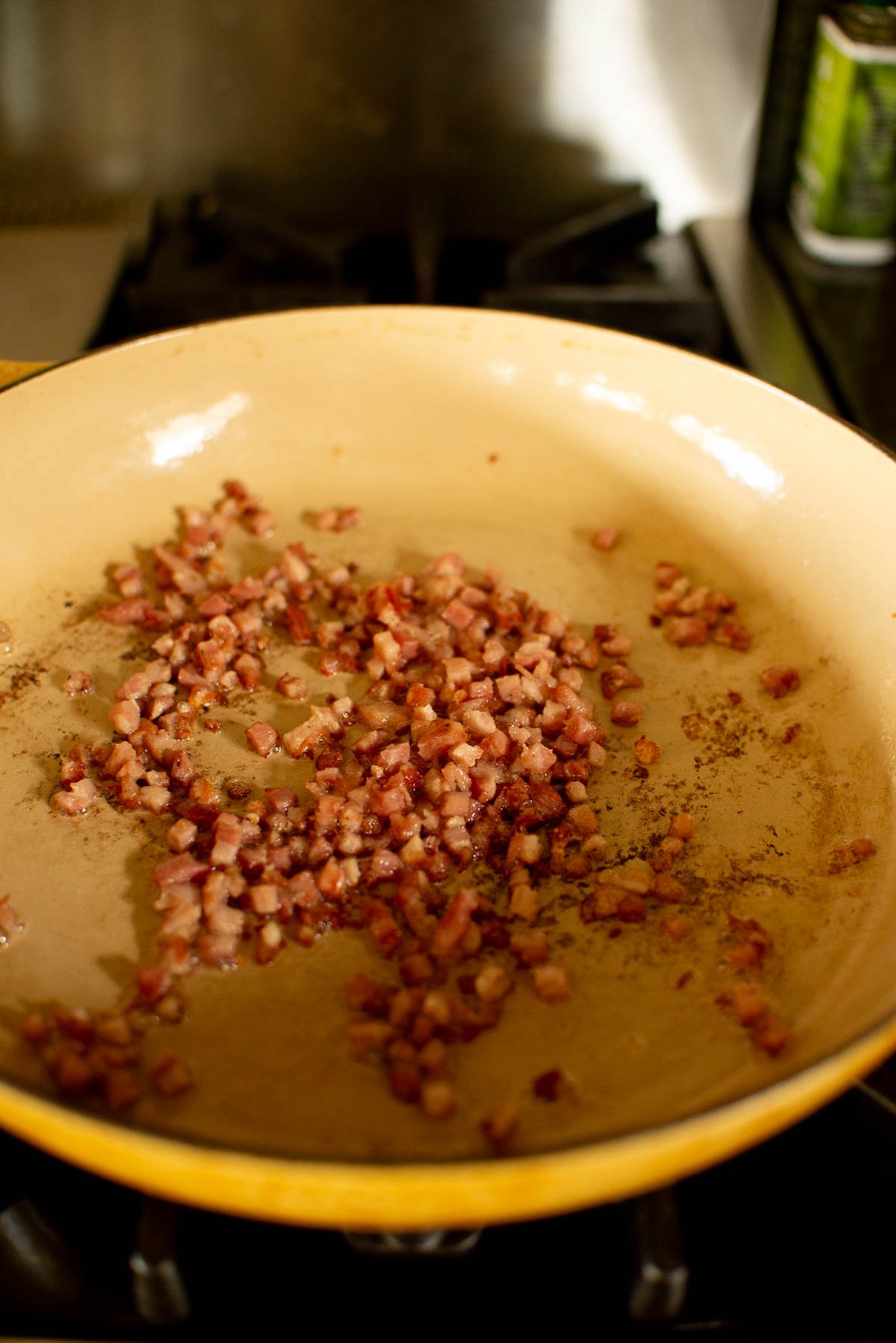 A large beige skillet with diced, browned pancetta on a stovetop.