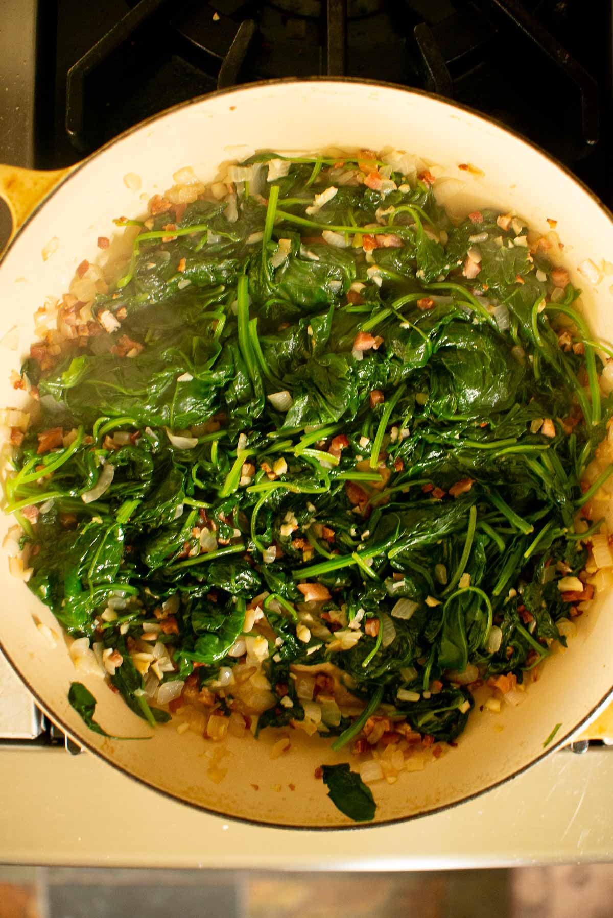 A skillet on the stove contains sautéed spinach mixed with chopped onions and small diced pieces of red and green vegetables.