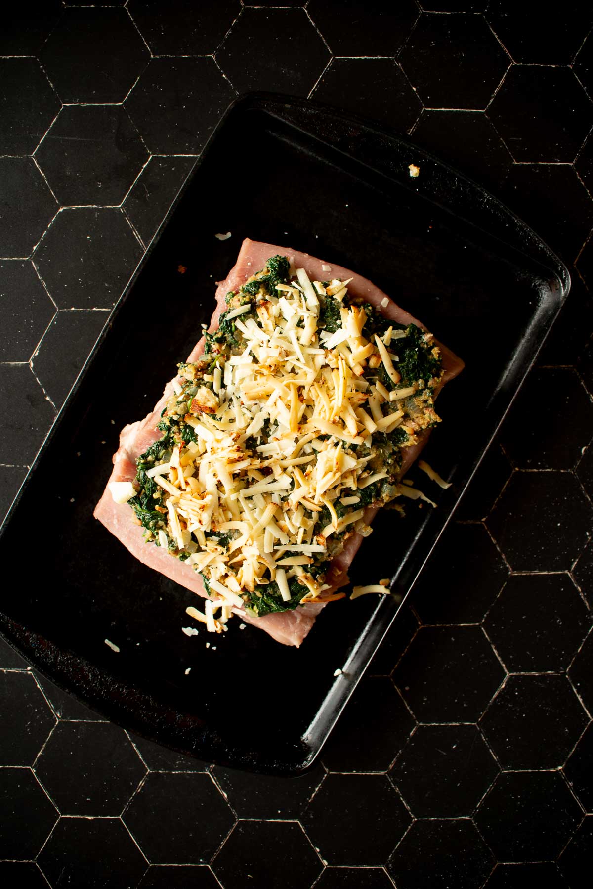 A raw piece of meat topped with spinach and shredded cheese sits on a black baking tray on a dark hexagonal-tiled surface.