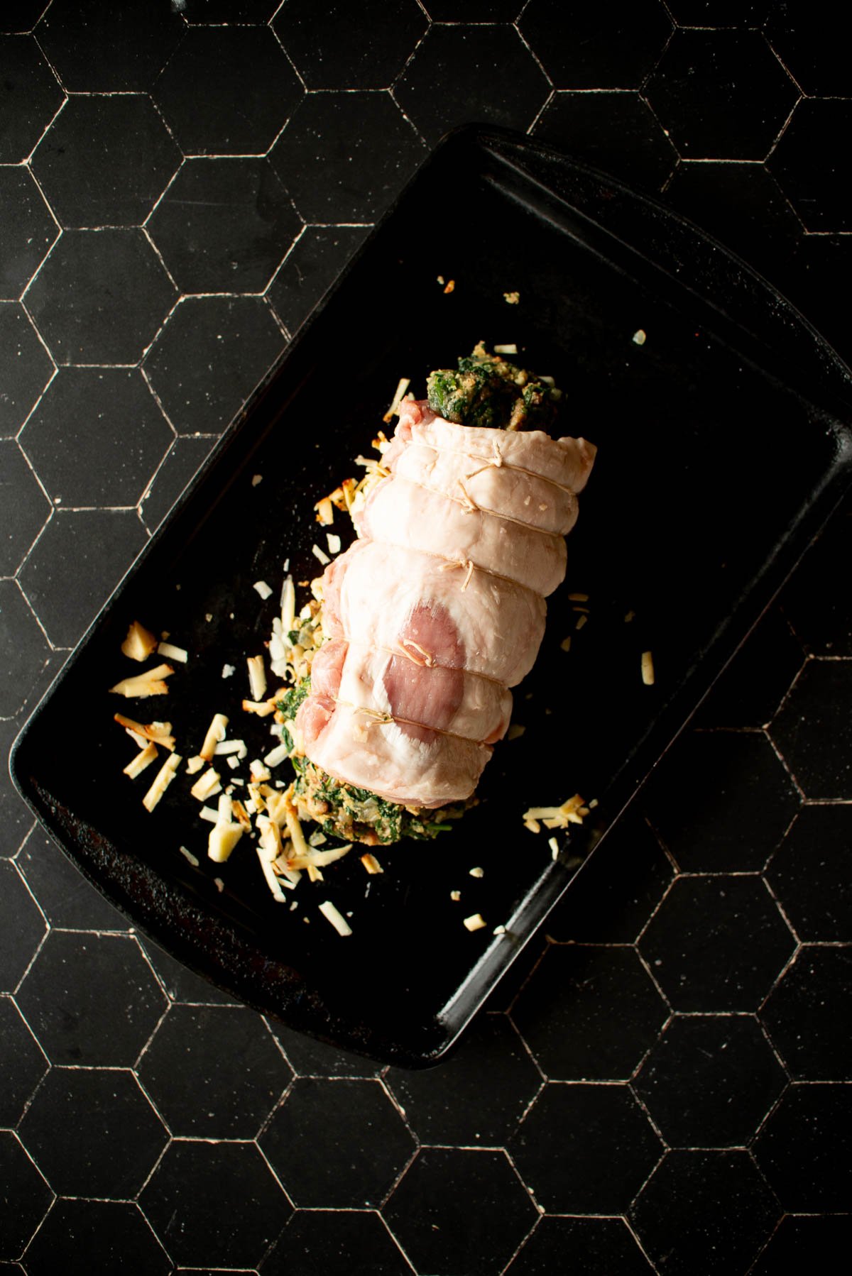 A stuffed and tied rolled meat roast is placed in a black baking dish, sprinkled with shredded cheese and herbs, set on a black hexagonal-patterned surface.