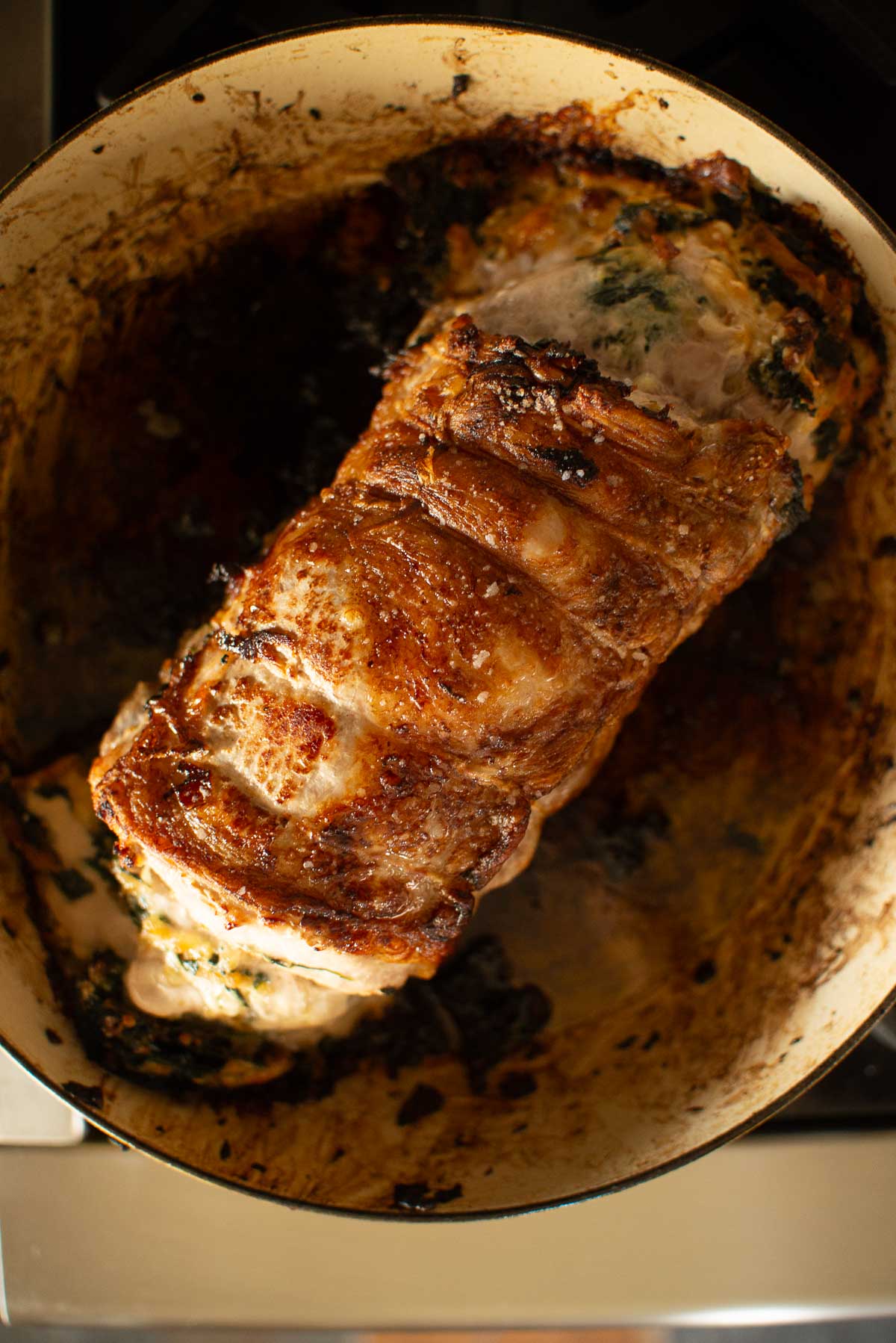 A roasted, browned stuffed pork loin sits in a cast iron skillet. The pork is tied with twine and appears stuffed with herbs. The skillet has charred remnants from the cooking process.