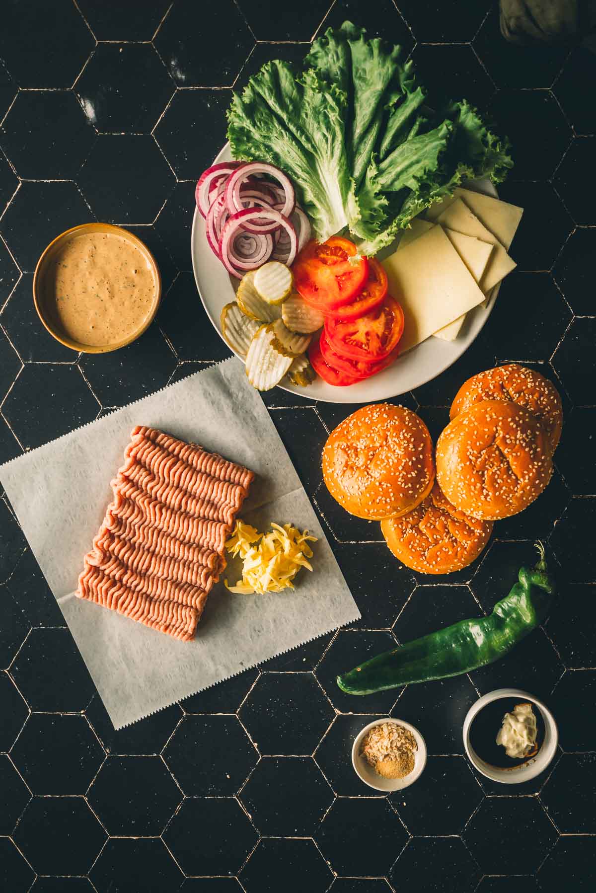 Ingredients for making turkey burgers arranged on a black tile surface. Includes ground meat, burger buns, lettuce, tomato, cheese, pickles, onions, condiments, and a green chili.