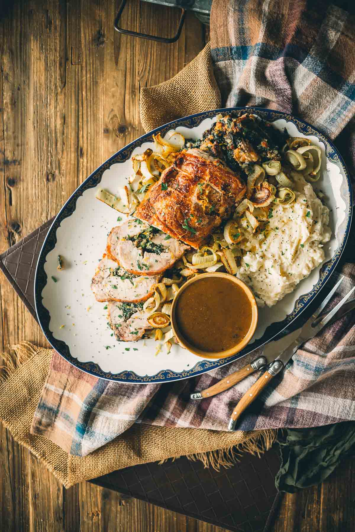 A platter with sliced stuffed pork loin, caramelized onions, mashed potatoes, stuffing, and a bowl of gravy, set on a wooden table with a cloth.