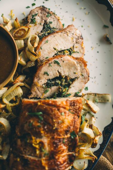 Close-up of sliced, stuffed pork tenderloin with herbs and leafy greens, garnished with chopped parsley and served alongside roasted leeks and a bowl of sauce.