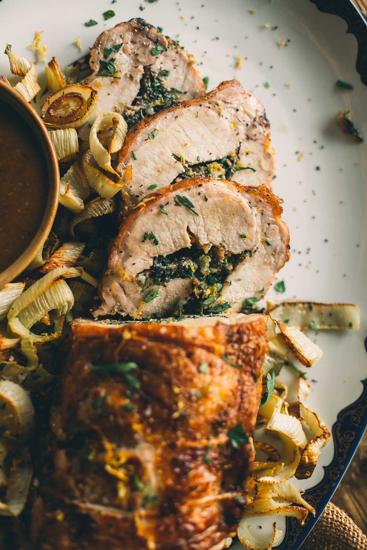 Close-up of sliced, stuffed pork tenderloin with herbs and leafy greens, garnished with chopped parsley and served alongside roasted leeks and a bowl of sauce.