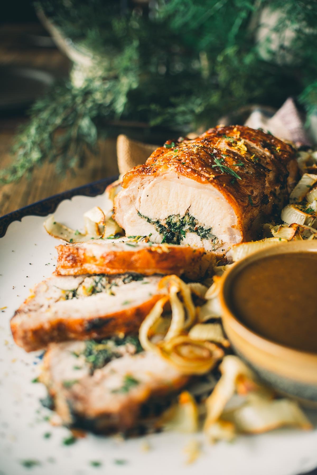 Plate with sliced stuffed pork loin garnished with herbs and onions, accompanied by a small bowl of gravy.