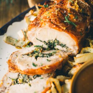 A cooked, sliced rolled pork roast stuffed with herbs and garnished with lemon zest and fresh herbs, served on a platter with caramelized onions.