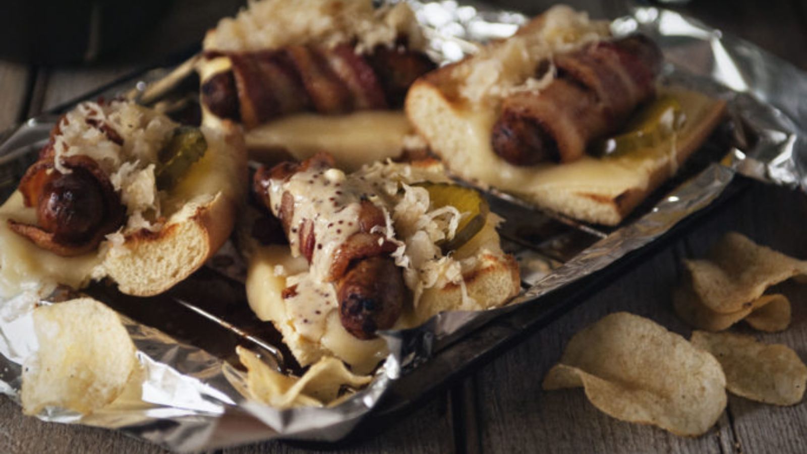 Bacon-wrapped brats on toasted rolls with sauerkraut, pickles, and mustard, served on a tray lined with foil, with potato chips on the side.