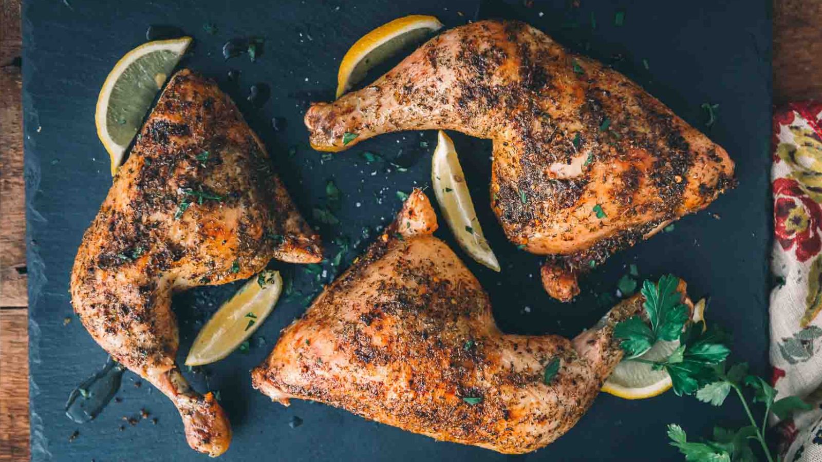 Three seasoned and roasted chicken legs are arranged on a slate board with lemon slices and sprigs of parsley.