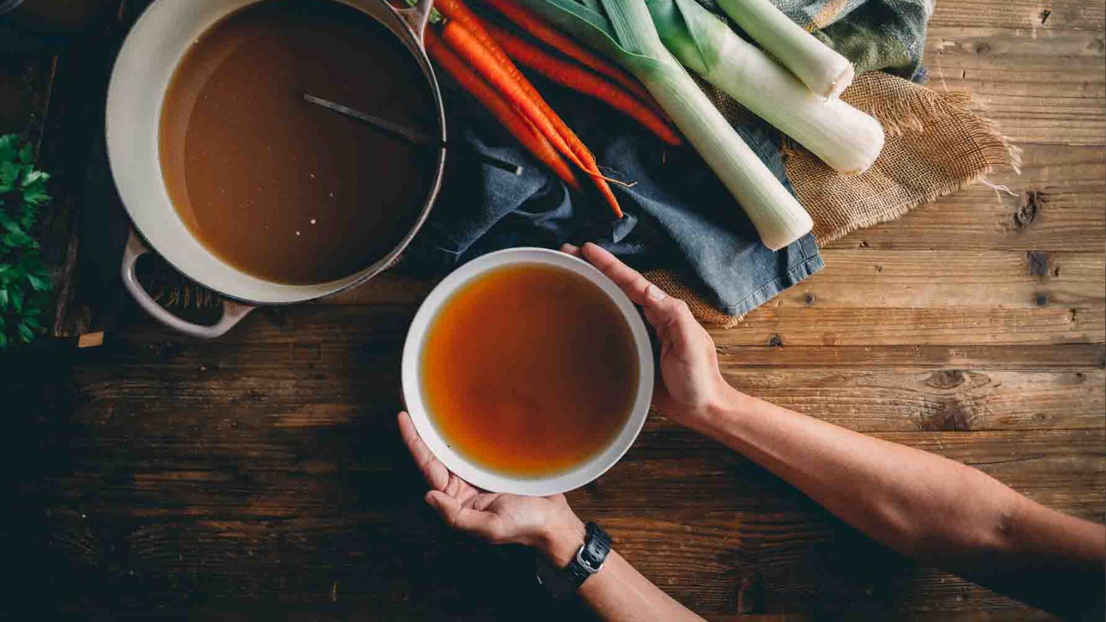 Person holding a bowl of broth over a wooden table with a pot of broth and vegetables, including carrots and leeks.