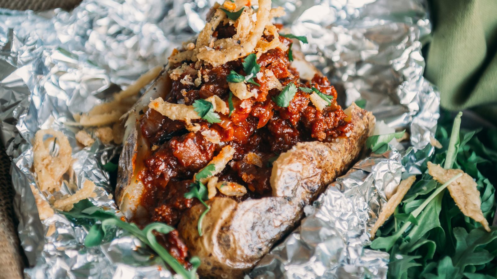 Baked potato topped with chili, crispy onions, and chopped herbs, wrapped in foil.