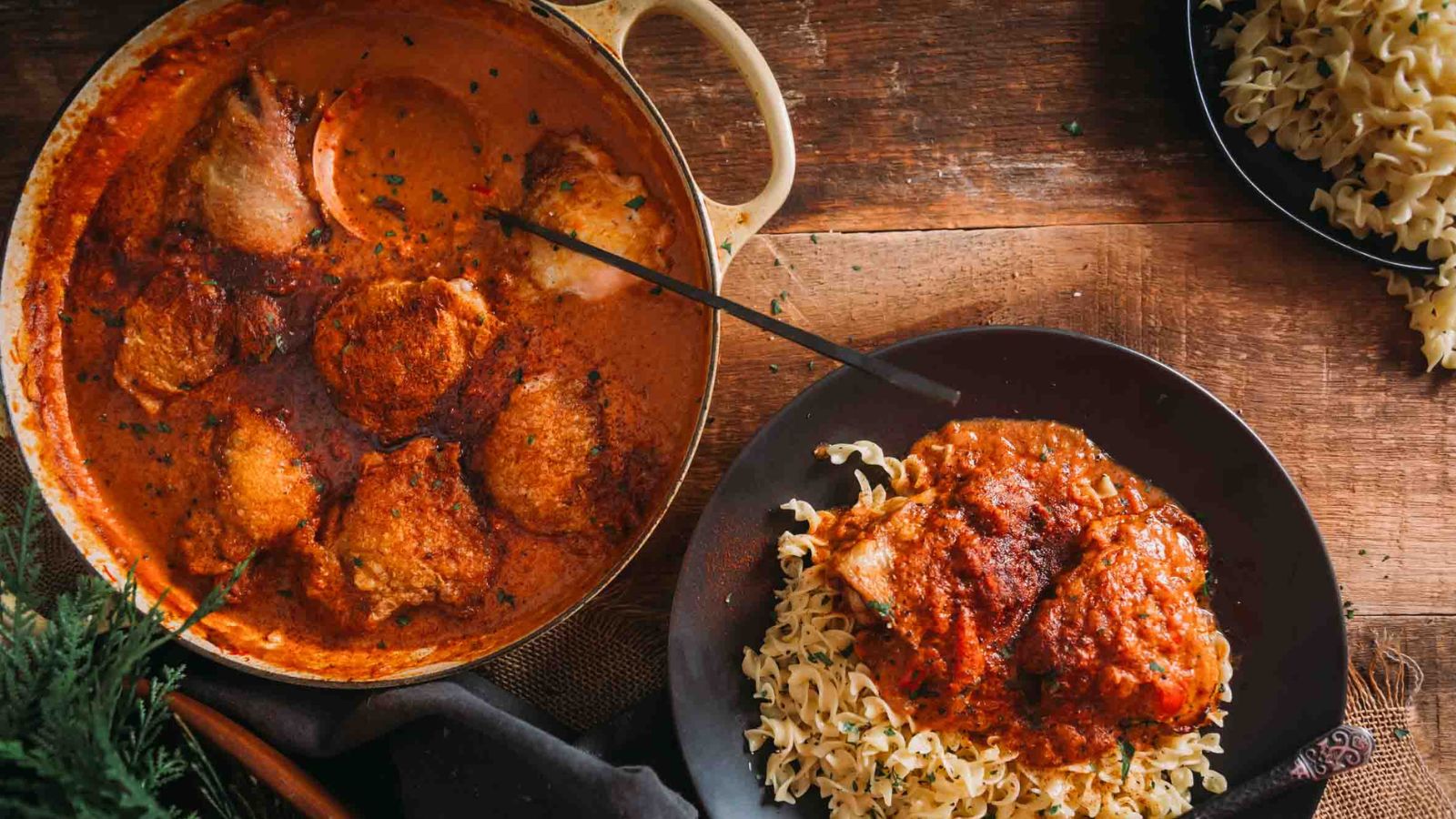 A pot of chicken in a creamy pepper sauce beside a plate of chicken served with pasta on a wooden table.