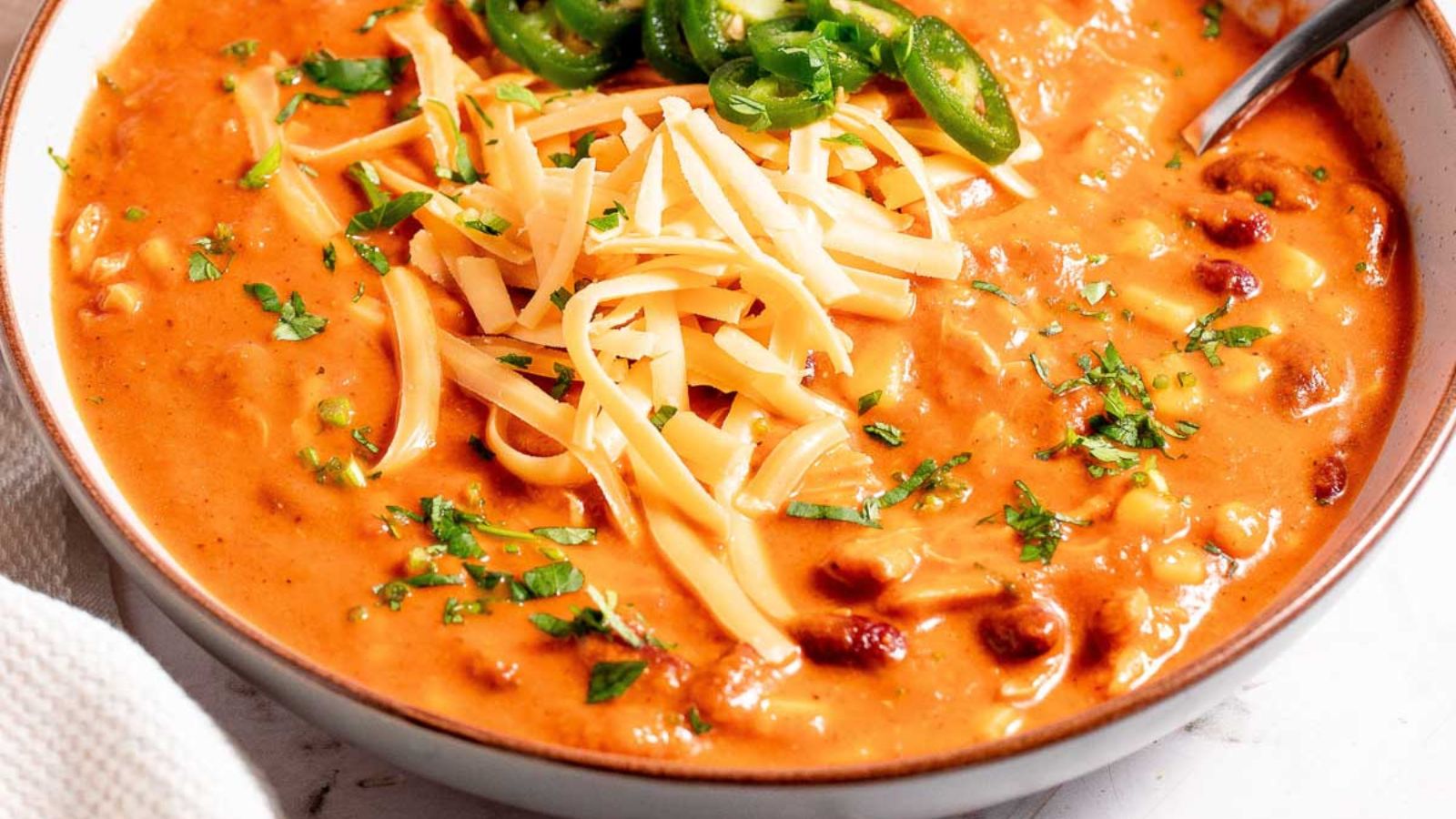 A bowl of creamy chili topped with shredded cheese, sliced jalapeños, and chopped cilantro.