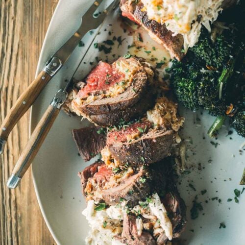 Sliced beef tenderloin stuffed with crab filling, served with baby broccoli on an oval white plate with a wooden-handled fork.