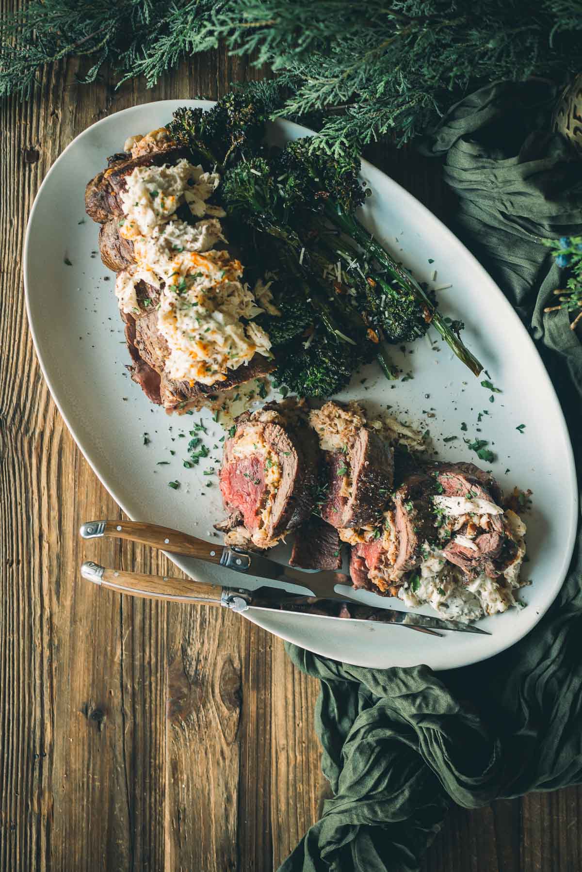 Sliced beef tenderloin stuffed with a creamy crab imperial on a white platter, garnished with broccoli rabe and parsley, placed on a rustic wooden table next to a green cloth.