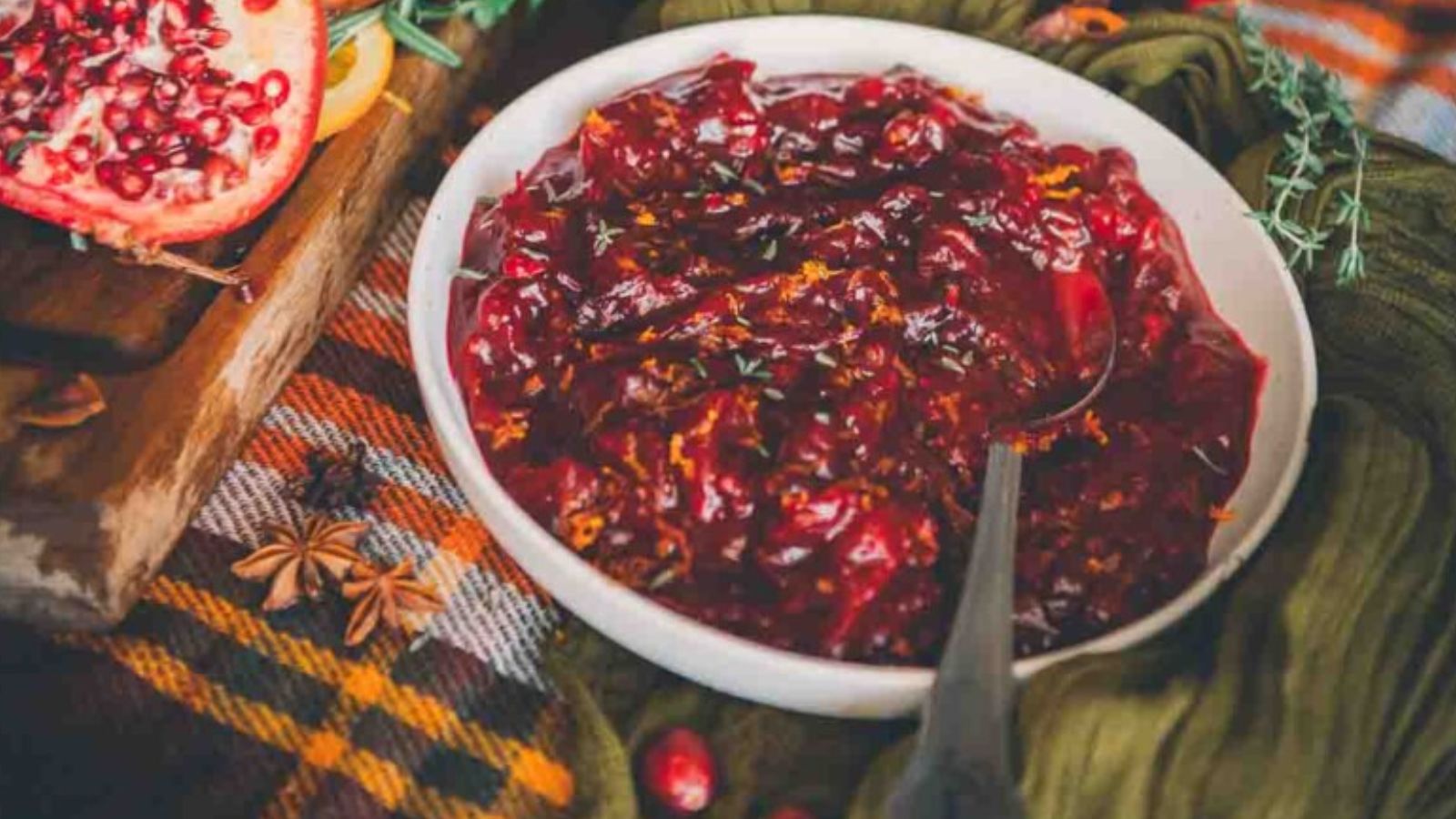 A white bowl filled with cranberry sauce on a plaid fabric background, accompanied by a halved pomegranate and star anise.