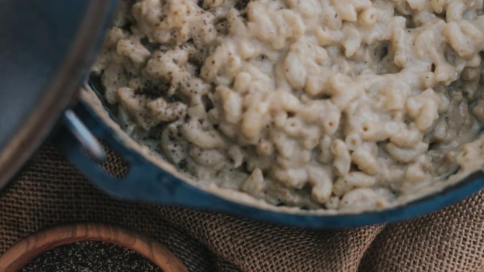 A pot of creamy macaroni and cheese seasoned with black pepper, placed on a burlap cloth near a wooden bowl.