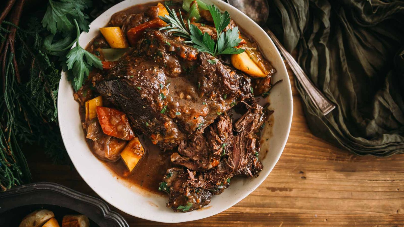 A bowl of pot roast with vegetables, garnished with parsley, on a wooden table.