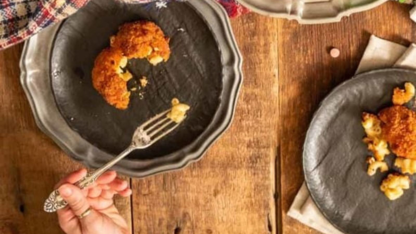 A hand holding a fork above a plate with a partially eaten croquette and scattered macaroni on a wooden table.