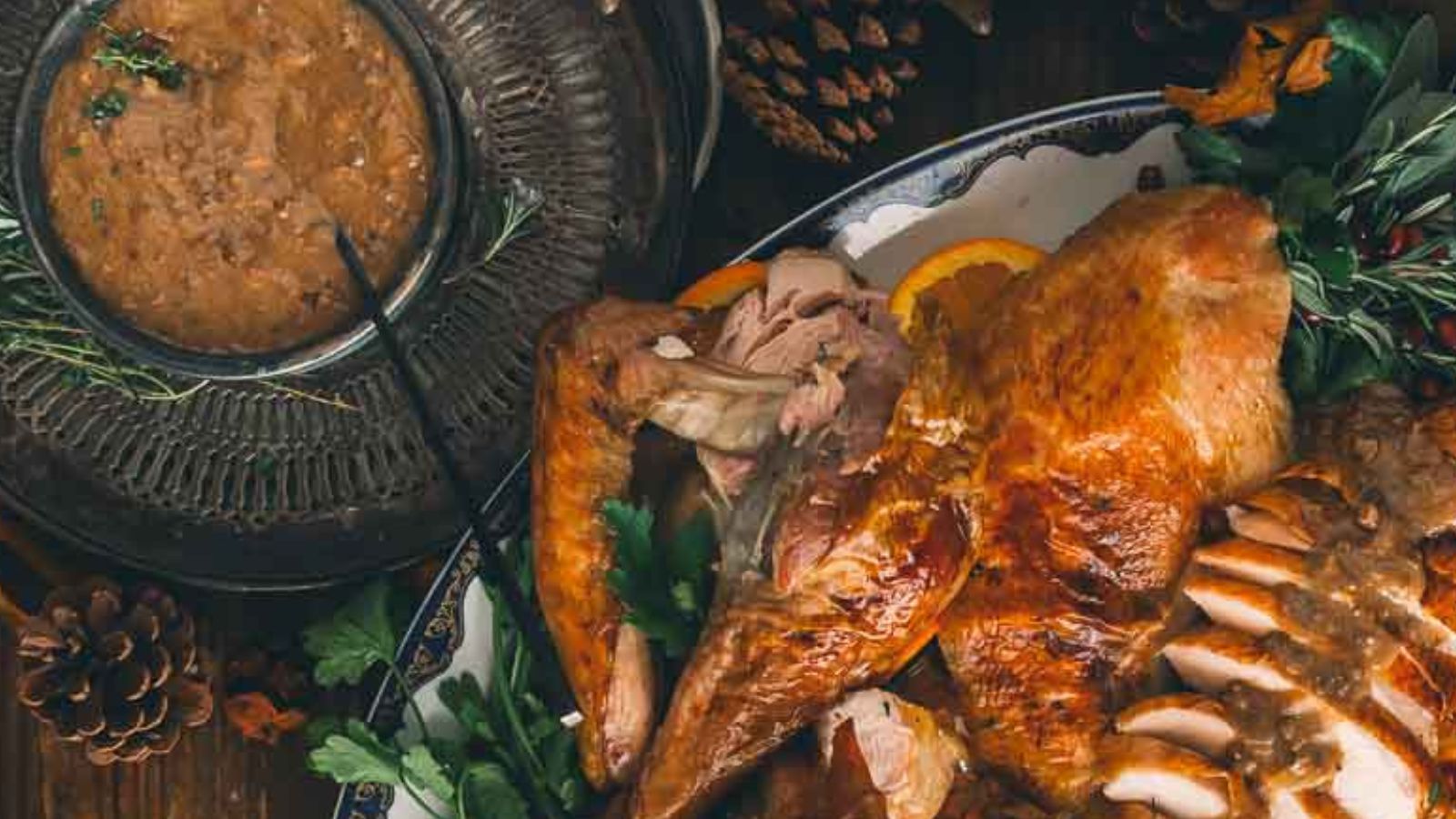 A roast turkey is served on a platter, garnished with herbs and sliced oranges. A bowl of gravy is nearby, surrounded by pinecones.