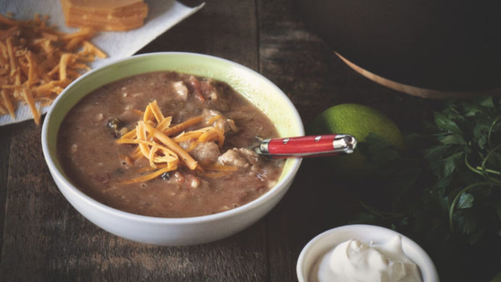 Bowl of soup with chunks of meat and vegetables, topped with shredded cheese. A red-handled spoon rests in the bowl. Surrounding items include shredded cheese, lime, herbs, and sour cream.