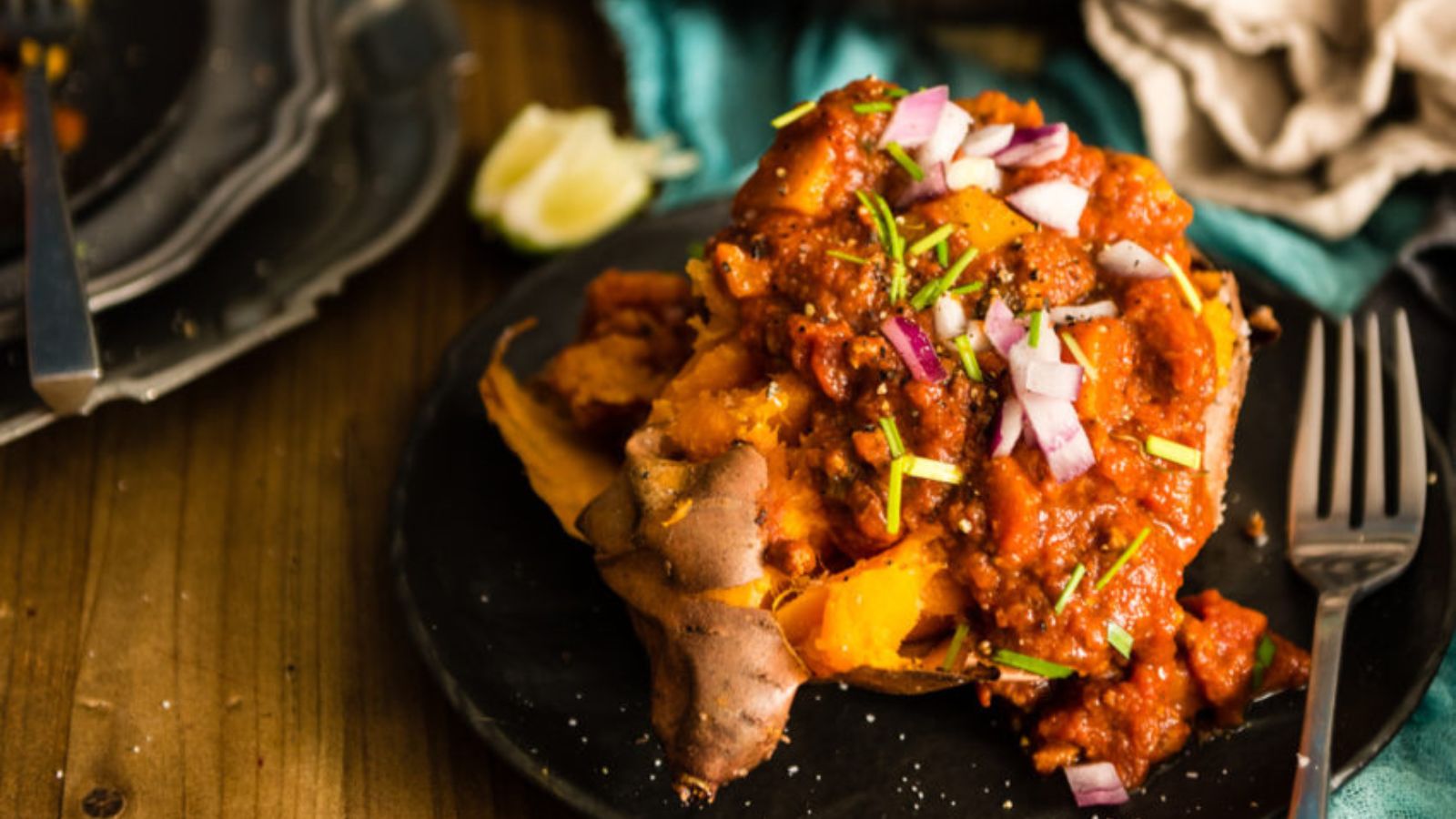 Baked sweet potato topped with chili, chopped red onions, and herbs on a black plate; lime wedge and cutlery nearby.